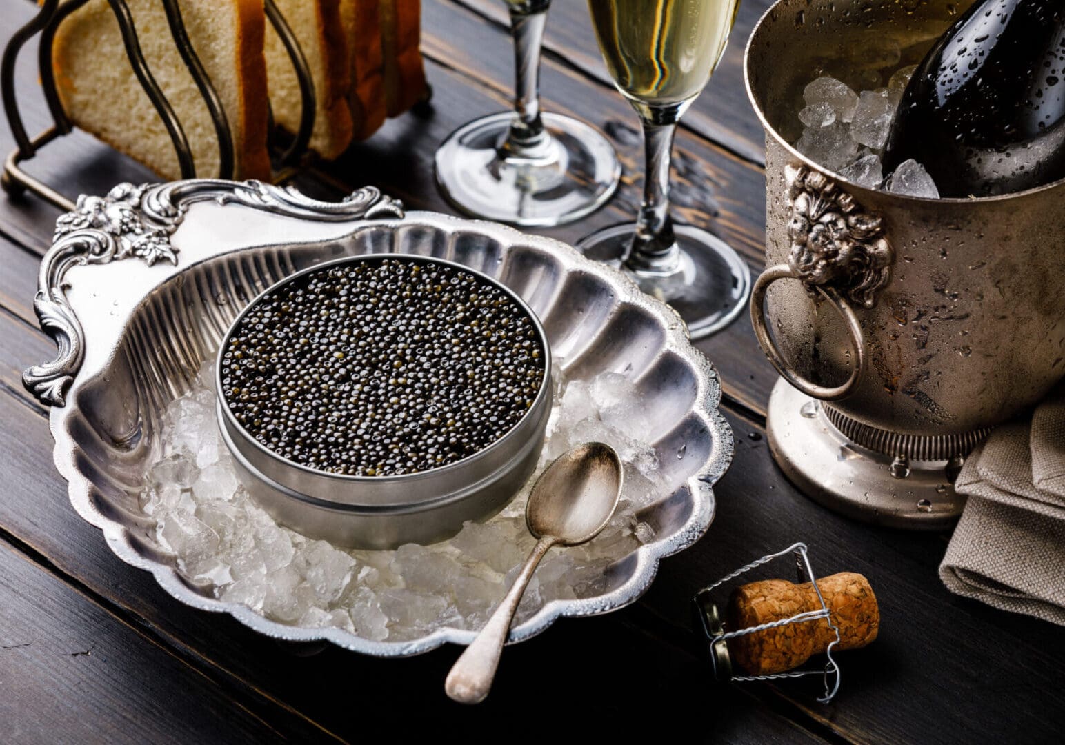 Black caviar in can on ice in silver bowl, bread and champagne in ice bucket on black wooden background